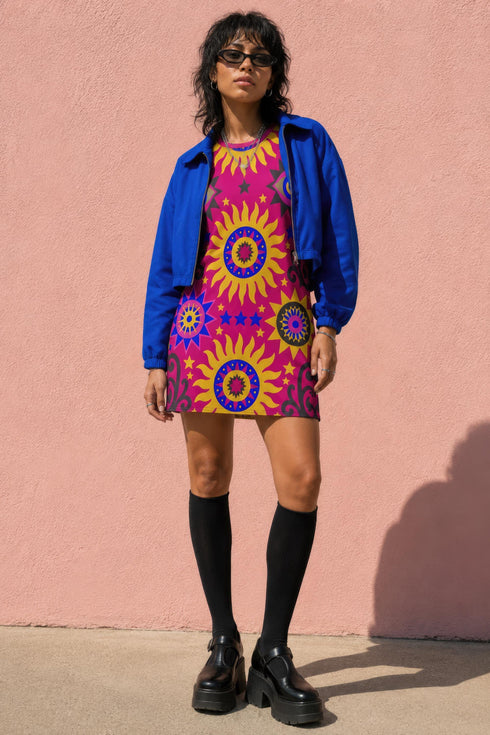 Short sleeve crew neck t-shirt dress in a relaxed, oversized straight cut, made of 100% polyester with a soft matte finish. An edge-to-edge all-over print shows yellow radial sunburst medallions with jagged rays on a hot pink background, layered blue and pink circular centers, rows of small blue stars, a partial eye near the top, and dark charcoal swirling vine filigree along the hem. Psychedelic 70s retro, celestial, whimsigoth maximalist alt fashion.