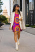 Short sleeve crew neck t-shirt dress in a relaxed, oversized straight cut, made of 100% polyester with a soft matte finish. An edge-to-edge all-over print shows yellow radial sunburst medallions with jagged rays on a hot pink background, layered blue and pink circular centers, rows of small blue stars, a partial eye near the top, and dark charcoal swirling vine filigree along the hem. Psychedelic 70s retro, celestial, whimsigoth maximalist alt fashion.