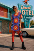 Short sleeve crew neck t-shirt dress in a relaxed, oversized straight cut, made of 100% polyester with a soft matte finish. An edge-to-edge all-over print on a medium purple background shows vertically stacked daisies with orange petals and yellow centers, scattered white four-point stars, and blue and green wavy side borders filled with small black and white daisies. Retro 70s floral, groovy maximalist alt fashion.