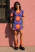 Short sleeve crew neck t-shirt dress in a relaxed, oversized straight cut, made of 100% polyester with a soft matte finish. An edge-to-edge all-over print on a medium purple background shows vertically stacked daisies with orange petals and yellow centers, scattered white four-point stars, and blue and green wavy side borders filled with small black and white daisies. Retro 70s floral, groovy maximalist alt fashion.