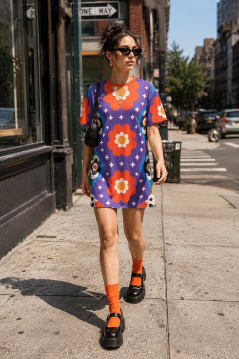 Short sleeve crew neck t-shirt dress in a relaxed, oversized straight cut, made of 100% polyester with a soft matte finish. An edge-to-edge all-over print on a medium purple background shows vertically stacked daisies with orange petals and yellow centers, scattered white four-point stars, and blue and green wavy side borders filled with small black and white daisies. Retro 70s floral, groovy maximalist alt fashion.