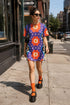 Short sleeve crew neck t-shirt dress in a relaxed, oversized straight cut, made of 100% polyester with a soft matte finish. An edge-to-edge all-over print on a medium purple background shows vertically stacked daisies with orange petals and yellow centers, scattered white four-point stars, and blue and green wavy side borders filled with small black and white daisies. Retro 70s floral, groovy maximalist alt fashion.