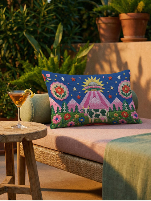 Rectangular 20 by 12 inch lumbar pillow cover printed to look like thick crewel embroidery on a deep royal blue night sky dotted with orange, yellow, and white stars. A silver, pink, and yellow flying saucer beams a bright pink cone of light onto a white cow with green spots standing in green grass with pine trees and red and pink flowers. Smooth flat 2D print, not raised stitching. Maximalist sci-fi decor for a bed, sofa, or reading nook.