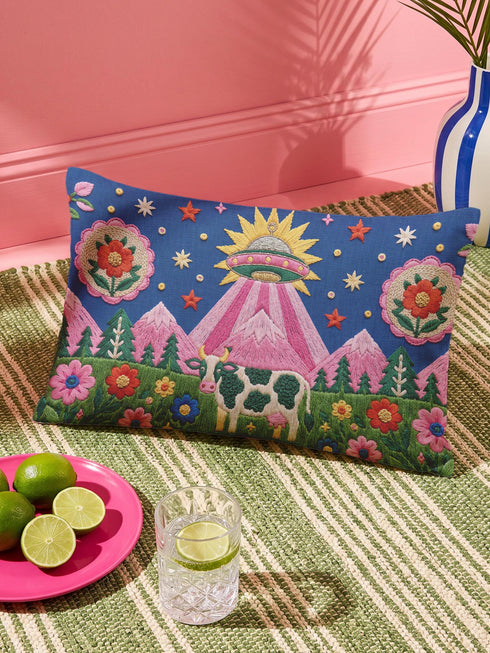 Rectangular 20 by 12 inch lumbar pillow cover printed to look like thick crewel embroidery on a deep royal blue night sky dotted with orange, yellow, and white stars. A silver, pink, and yellow flying saucer beams a bright pink cone of light onto a white cow with green spots standing in green grass with pine trees and red and pink flowers. Smooth flat 2D print, not raised stitching. Maximalist sci-fi decor for a bed, sofa, or reading nook.