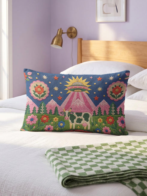 Rectangular 20 by 12 inch lumbar pillow cover printed to look like thick crewel embroidery on a deep royal blue night sky dotted with orange, yellow, and white stars. A silver, pink, and yellow flying saucer beams a bright pink cone of light onto a white cow with green spots standing in green grass with pine trees and red and pink flowers. Smooth flat 2D print, not raised stitching. Maximalist sci-fi decor for a bed, sofa, or reading nook.