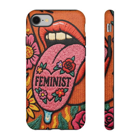 Dual layer matte phone case with a 2D print that mimics knit and embroidery texture. A retro open mouth with its tongue out sits over a pink heart stitched with the word FEMINIST in black letters, on an orange knit-look background filled with pink flowers, yellow sunflowers, red roses, purple and orange blooms, teal leaves, and a turquoise drip, all outlined in bold black. Smooth printed surface, open ports, camera cutout. Feminist maximalist phone accessory.