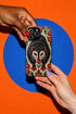 Dual layer matte phone case with a 2D print that mimics thick crewel embroidery. An angry black possum with a white face, bared sharp teeth, and bright red eyes and claws sits at the center on a cream background, framed by a symmetrical border of red folk art daisies with yellow centers and green leaves. Smooth printed surface with no raised threads, open ports, and a precise camera cutout. Goblincore, creepy cute, maximalist phone accessory.
