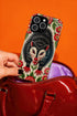 Dual layer matte phone case with a 2D print that mimics thick crewel embroidery. An angry black possum with a white face, bared sharp teeth, and bright red eyes and claws sits at the center on a cream background, framed by a symmetrical border of red folk art daisies with yellow centers and green leaves. Smooth printed surface with no raised threads, open ports, and a precise camera cutout. Goblincore, creepy cute, maximalist phone accessory.