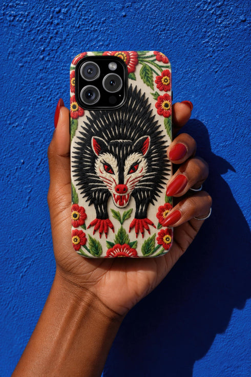 Dual layer matte phone case with a 2D print that mimics thick crewel embroidery. An angry black possum with a white face, bared sharp teeth, and bright red eyes and claws sits at the center on a cream background, framed by a symmetrical border of red folk art daisies with yellow centers and green leaves. Smooth printed surface with no raised threads, open ports, and a precise camera cutout. Goblincore, creepy cute, maximalist phone accessory.
