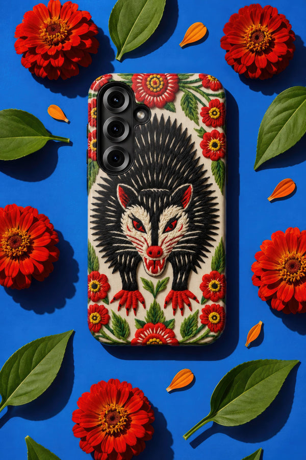 Dual layer matte phone case with a 2D print that mimics thick crewel embroidery. An angry black possum with a white face, bared sharp teeth, and bright red eyes and claws sits at the center on a cream background, framed by a symmetrical border of red folk art daisies with yellow centers and green leaves. Smooth printed surface with no raised threads, open ports, and a precise camera cutout. Goblincore, creepy cute, maximalist phone accessory.