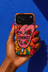 Dual layer matte phone case with a 2D print that mimics knit and embroidery texture. A retro open mouth with its tongue out sits over a pink heart stitched with the word FEMINIST in black letters, on an orange knit-look background filled with pink flowers, yellow sunflowers, red roses, purple and orange blooms, teal leaves, and a turquoise drip, all outlined in bold black. Smooth printed surface, open ports, camera cutout. Feminist maximalist phone accessory.