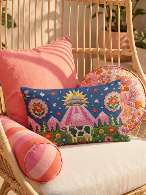 Rectangular 20 by 12 inch lumbar pillow cover printed to look like thick crewel embroidery on a deep royal blue night sky dotted with orange, yellow, and white stars. A silver, pink, and yellow flying saucer beams a bright pink cone of light onto a white cow with green spots standing in green grass with pine trees and red and pink flowers. Smooth flat 2D print, not raised stitching. Maximalist sci-fi decor for a bed, sofa, or reading nook.