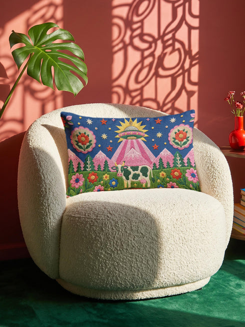 Rectangular 20 by 12 inch lumbar pillow cover printed to look like thick crewel embroidery on a deep royal blue night sky dotted with orange, yellow, and white stars. A silver, pink, and yellow flying saucer beams a bright pink cone of light onto a white cow with green spots standing in green grass with pine trees and red and pink flowers. Smooth flat 2D print, not raised stitching. Maximalist sci-fi decor for a bed, sofa, or reading nook.