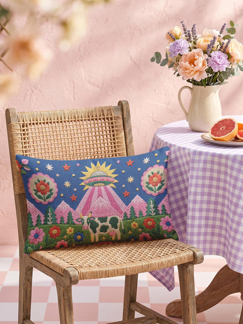 Rectangular 20 by 12 inch lumbar pillow cover printed to look like thick crewel embroidery on a deep royal blue night sky dotted with orange, yellow, and white stars. A silver, pink, and yellow flying saucer beams a bright pink cone of light onto a white cow with green spots standing in green grass with pine trees and red and pink flowers. Smooth flat 2D print, not raised stitching. Maximalist sci-fi decor for a bed, sofa, or reading nook.
