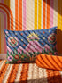 Rectangular 20 by 12 inch lumbar pillow cover printed to look like thick crewel embroidery on a deep royal blue night sky dotted with orange, yellow, and white stars. A silver, pink, and yellow flying saucer beams a bright pink cone of light onto a white cow with green spots standing in green grass with pine trees and red and pink flowers. Smooth flat 2D print, not raised stitching. Maximalist sci-fi decor for a bed, sofa, or reading nook.
