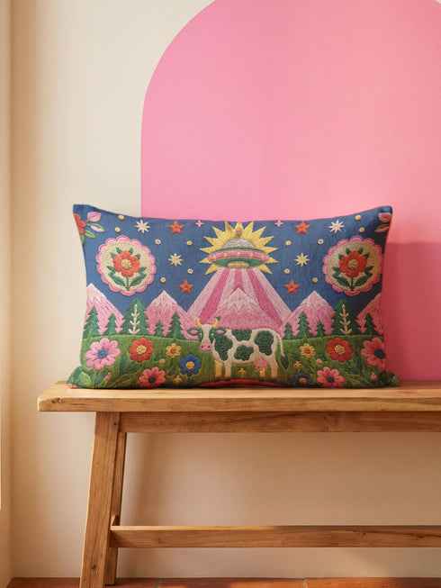 Rectangular 20 by 12 inch lumbar pillow cover printed to look like thick crewel embroidery on a deep royal blue night sky dotted with orange, yellow, and white stars. A silver, pink, and yellow flying saucer beams a bright pink cone of light onto a white cow with green spots standing in green grass with pine trees and red and pink flowers. Smooth flat 2D print, not raised stitching. Maximalist sci-fi decor for a bed, sofa, or reading nook.