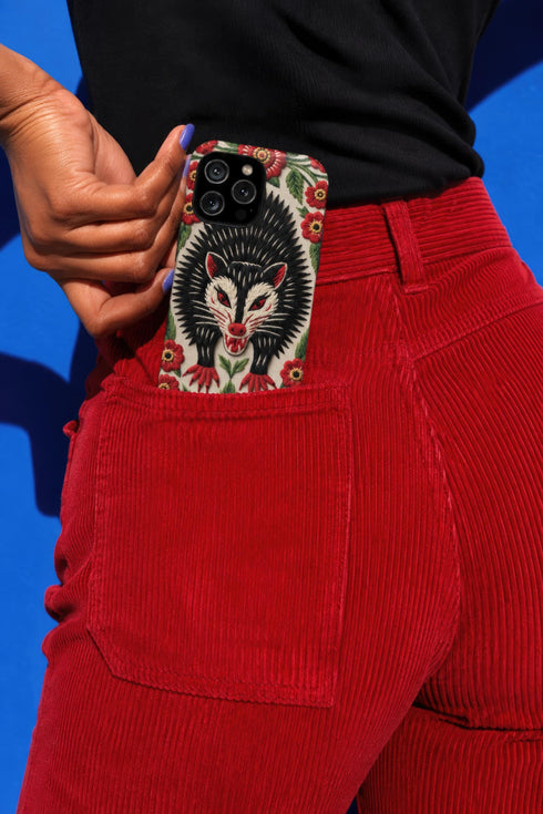 Dual layer matte phone case with a 2D print that mimics thick crewel embroidery. An angry black possum with a white face, bared sharp teeth, and bright red eyes and claws sits at the center on a cream background, framed by a symmetrical border of red folk art daisies with yellow centers and green leaves. Smooth printed surface with no raised threads, open ports, and a precise camera cutout. Goblincore, creepy cute, maximalist phone accessory.