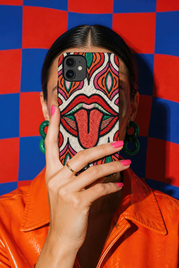 Dual layer matte phone case with a 2D print that mimics heavy crochet and thick knit yarn. A large open mouth with bright red lips and a red tongue sticking out sits at the center of a cream background, surrounded by mirrored teardrop and flame shapes in bright orange, dark green, and hot pink with black stitch-style outlines. Smooth printed surface with no raised threads, open ports, and camera cutout. Psychedelic 70s pop art phone accessory.