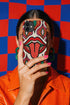 Dual layer matte phone case with a 2D print that mimics heavy crochet and thick knit yarn. A large open mouth with bright red lips and a red tongue sticking out sits at the center of a cream background, surrounded by mirrored teardrop and flame shapes in bright orange, dark green, and hot pink with black stitch-style outlines. Smooth printed surface with no raised threads, open ports, and camera cutout. Psychedelic 70s pop art phone accessory.