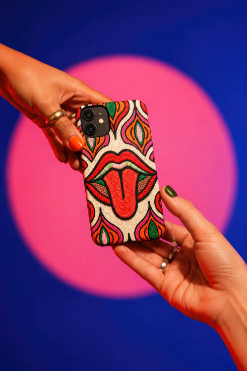 Dual layer matte phone case with a 2D print that mimics heavy crochet and thick knit yarn. A large open mouth with bright red lips and a red tongue sticking out sits at the center of a cream background, surrounded by mirrored teardrop and flame shapes in bright orange, dark green, and hot pink with black stitch-style outlines. Smooth printed surface with no raised threads, open ports, and camera cutout. Psychedelic 70s pop art phone accessory.