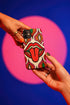 Dual layer matte phone case with a 2D print that mimics heavy crochet and thick knit yarn. A large open mouth with bright red lips and a red tongue sticking out sits at the center of a cream background, surrounded by mirrored teardrop and flame shapes in bright orange, dark green, and hot pink with black stitch-style outlines. Smooth printed surface with no raised threads, open ports, and camera cutout. Psychedelic 70s pop art phone accessory.