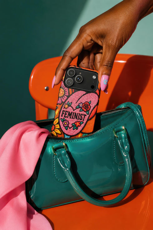 Dual layer matte phone case with a 2D print that mimics knit and embroidery texture. A retro open mouth with its tongue out sits over a pink heart stitched with the word FEMINIST in black letters, on an orange knit-look background filled with pink flowers, yellow sunflowers, red roses, purple and orange blooms, teal leaves, and a turquoise drip, all outlined in bold black. Smooth printed surface, open ports, camera cutout. Feminist maximalist phone accessory.