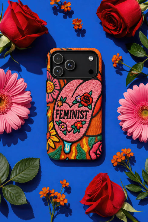 Dual layer matte phone case with a 2D print that mimics knit and embroidery texture. A retro open mouth with its tongue out sits over a pink heart stitched with the word FEMINIST in black letters, on an orange knit-look background filled with pink flowers, yellow sunflowers, red roses, purple and orange blooms, teal leaves, and a turquoise drip, all outlined in bold black. Smooth printed surface, open ports, camera cutout. Feminist maximalist phone accessory.