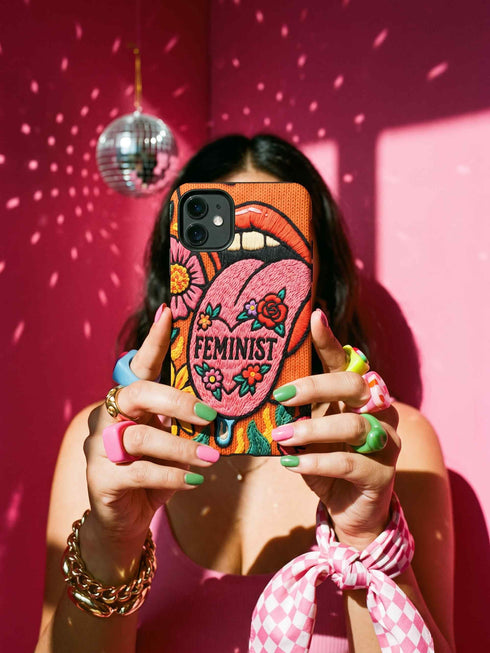 Dual layer matte phone case with a 2D print that mimics knit and embroidery texture. A retro open mouth with its tongue out sits over a pink heart stitched with the word FEMINIST in black letters, on an orange knit-look background filled with pink flowers, yellow sunflowers, red roses, purple and orange blooms, teal leaves, and a turquoise drip, all outlined in bold black. Smooth printed surface, open ports, camera cutout. Feminist maximalist phone accessory.