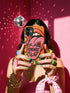 Dual layer matte phone case with a 2D print that mimics knit and embroidery texture. A retro open mouth with its tongue out sits over a pink heart stitched with the word FEMINIST in black letters, on an orange knit-look background filled with pink flowers, yellow sunflowers, red roses, purple and orange blooms, teal leaves, and a turquoise drip, all outlined in bold black. Smooth printed surface, open ports, camera cutout. Feminist maximalist phone accessory.