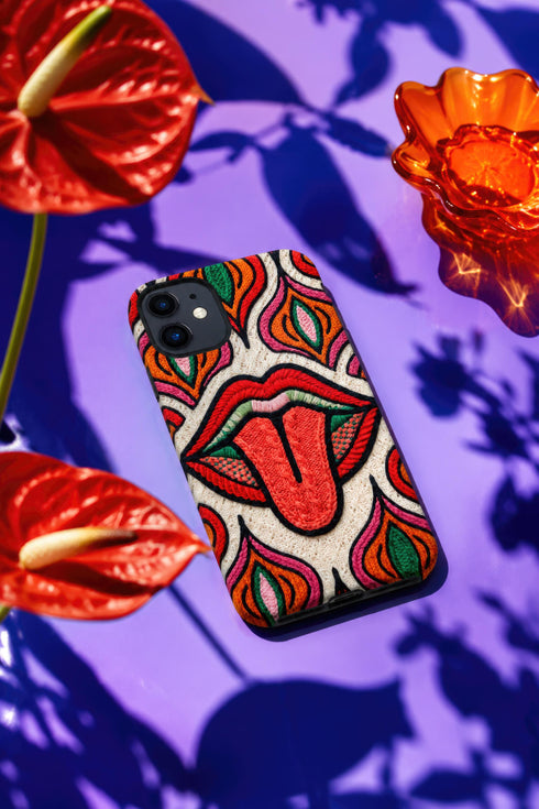 Dual layer matte phone case with a 2D print that mimics heavy crochet and thick knit yarn. A large open mouth with bright red lips and a red tongue sticking out sits at the center of a cream background, surrounded by mirrored teardrop and flame shapes in bright orange, dark green, and hot pink with black stitch-style outlines. Smooth printed surface with no raised threads, open ports, and camera cutout. Psychedelic 70s pop art phone accessory.