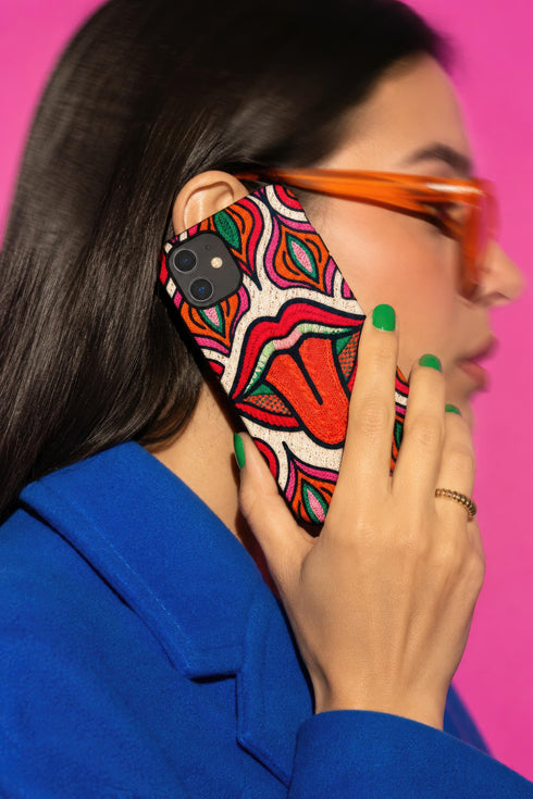 Dual layer matte phone case with a 2D print that mimics heavy crochet and thick knit yarn. A large open mouth with bright red lips and a red tongue sticking out sits at the center of a cream background, surrounded by mirrored teardrop and flame shapes in bright orange, dark green, and hot pink with black stitch-style outlines. Smooth printed surface with no raised threads, open ports, and camera cutout. Psychedelic 70s pop art phone accessory.