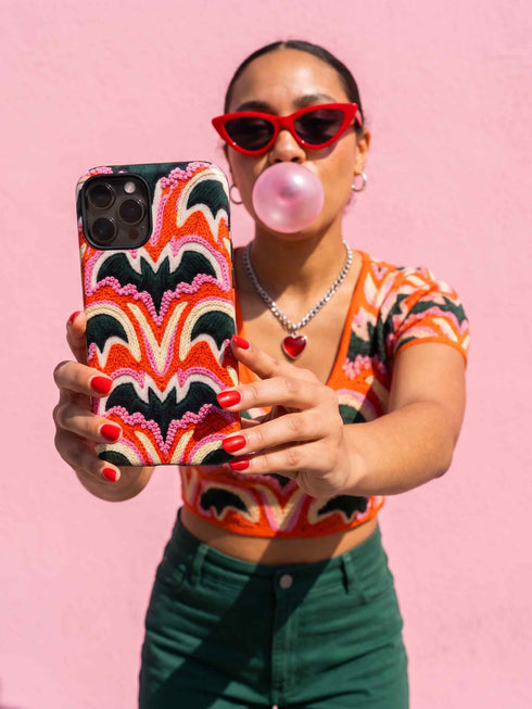 Dual layer matte phone case with a 2D print that mimics thick, heavy crochet yarn. Rows of stacked black bats repeat across a vibrant rust-orange background, accented with bright pink faux yarn bobbles and framed by cream arched lines. Smooth printed surface with no raised threads, open ports, and a precise camera cutout. Pastel goth, creepy cute, Summerween and Halloween phone accessory with a cozy handmade look.