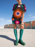 Short sleeve crew neck t-shirt dress in a relaxed, oversized straight cut, made of 100% polyester with a soft matte finish. An edge-to-edge all-over print on a true black background shows a large blue and white evil eye inside an orange wavy sunburst with a white jagged border, pink daisies with orange centers, white four-petal stars, and wavy blue and teal green leaves. Psychedelic whimsigoth, witchy celestial maximalist alt fashion.