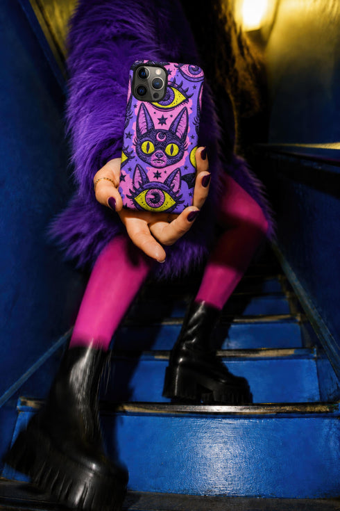 Dual layer matte phone case with a 2D print that mimics raised embroidery. Dark purple vampire bat cat heads with tall pointed ears, glowing chartreuse eyes, tiny fangs, and a pink crescent moon on the forehead repeat across a mottled purple and hot pink background, scattered with large chartreuse evil eyes with purple irises and small dark stars. Smooth printed surface with no raised threads, open ports, and camera cutout. Pastel goth, whimsigoth, creepy cute phone accessory.