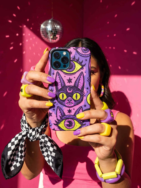 Dual layer matte phone case with a 2D print that mimics raised embroidery. Dark purple vampire bat cat heads with tall pointed ears, glowing chartreuse eyes, tiny fangs, and a pink crescent moon on the forehead repeat across a mottled purple and hot pink background, scattered with large chartreuse evil eyes with purple irises and small dark stars. Smooth printed surface with no raised threads, open ports, and camera cutout. Pastel goth, whimsigoth, creepy cute phone accessory.