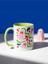 Glossy white ceramic 11 oz coffee mug with a lime green rim, inside, and handle and a full wraparound pastel goth print. Pink ghosts with paired red oval eyes and rounded scalloped lower edges float between wavy green and red ribbons. Blue starbursts, curved blue shapes, red teardrops, small colorful dots, and green diamond sparkles fill the white background. Creepy cute Halloween coffee mug with a playful, retro maximalist feel.