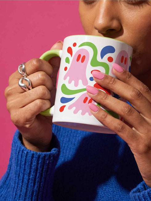 Glossy white ceramic 11 oz coffee mug with a lime green rim, inside, and handle and a full wraparound pastel goth print. Pink ghosts with paired red oval eyes and rounded scalloped lower edges float between wavy green and red ribbons. Blue starbursts, curved blue shapes, red teardrops, small colorful dots, and green diamond sparkles fill the white background. Creepy cute Halloween coffee mug with a playful, retro maximalist feel.