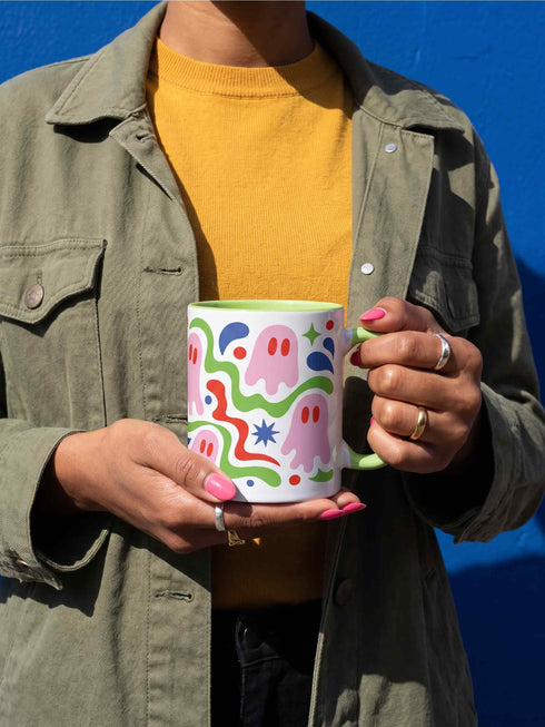 Glossy white ceramic 11 oz coffee mug with a lime green rim, inside, and handle and a full wraparound pastel goth print. Pink ghosts with paired red oval eyes and rounded scalloped lower edges float between wavy green and red ribbons. Blue starbursts, curved blue shapes, red teardrops, small colorful dots, and green diamond sparkles fill the white background. Creepy cute Halloween coffee mug with a playful, retro maximalist feel.