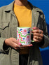 Glossy white ceramic 11 oz coffee mug with a lime green rim, inside, and handle and a full wraparound pastel goth print. Pink ghosts with paired red oval eyes and rounded scalloped lower edges float between wavy green and red ribbons. Blue starbursts, curved blue shapes, red teardrops, small colorful dots, and green diamond sparkles fill the white background. Creepy cute Halloween coffee mug with a playful, retro maximalist feel.
