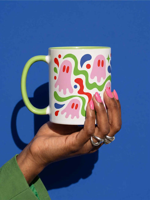 Glossy white ceramic 11 oz coffee mug with a lime green rim, inside, and handle and a full wraparound pastel goth print. Pink ghosts with paired red oval eyes and rounded scalloped lower edges float between wavy green and red ribbons. Blue starbursts, curved blue shapes, red teardrops, small colorful dots, and green diamond sparkles fill the white background. Creepy cute Halloween coffee mug with a playful, retro maximalist feel.