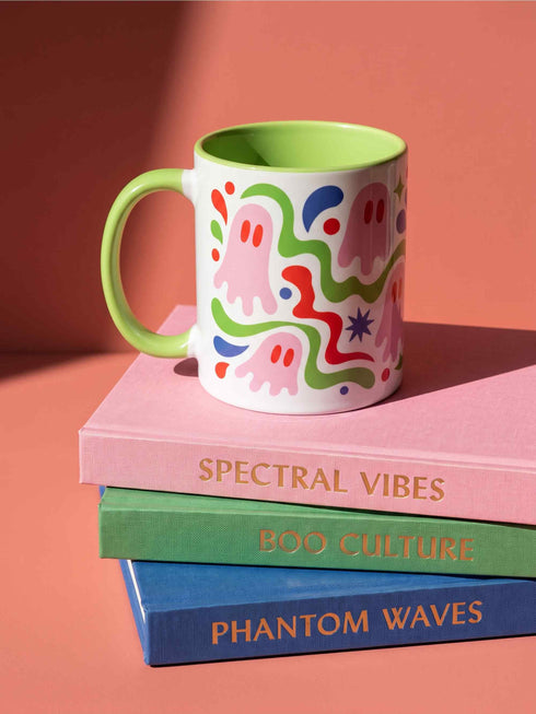 Glossy white ceramic 11 oz coffee mug with a lime green rim, inside, and handle and a full wraparound pastel goth print. Pink ghosts with paired red oval eyes and rounded scalloped lower edges float between wavy green and red ribbons. Blue starbursts, curved blue shapes, red teardrops, small colorful dots, and green diamond sparkles fill the white background. Creepy cute Halloween coffee mug with a playful, retro maximalist feel.
