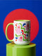 Glossy white ceramic 11 oz coffee mug with a lime green rim, inside, and handle and a full wraparound pastel goth print. Pink ghosts with paired red oval eyes and rounded scalloped lower edges float between wavy green and red ribbons. Blue starbursts, curved blue shapes, red teardrops, small colorful dots, and green diamond sparkles fill the white background. Creepy cute Halloween coffee mug with a playful, retro maximalist feel.