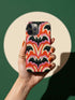 Dual layer matte phone case with a 2D print that mimics thick, heavy crochet yarn. Rows of stacked black bats repeat across a vibrant rust-orange background, accented with bright pink faux yarn bobbles and framed by cream arched lines. Smooth printed surface with no raised threads, open ports, and a precise camera cutout. Pastel goth, creepy cute, Summerween and Halloween phone accessory with a cozy handmade look.