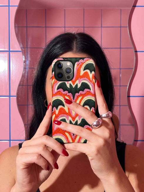 Dual layer matte phone case with a 2D print that mimics thick, heavy crochet yarn. Rows of stacked black bats repeat across a vibrant rust-orange background, accented with bright pink faux yarn bobbles and framed by cream arched lines. Smooth printed surface with no raised threads, open ports, and a precise camera cutout. Pastel goth, creepy cute, Summerween and Halloween phone accessory with a cozy handmade look.