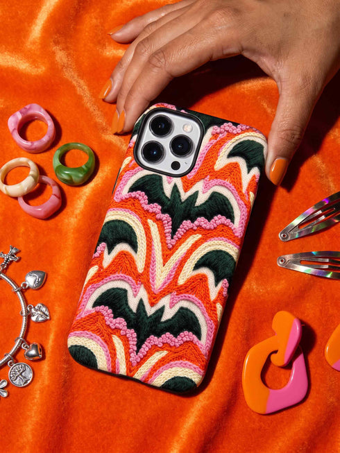 Dual layer matte phone case with a 2D print that mimics thick, heavy crochet yarn. Rows of stacked black bats repeat across a vibrant rust-orange background, accented with bright pink faux yarn bobbles and framed by cream arched lines. Smooth printed surface with no raised threads, open ports, and a precise camera cutout. Pastel goth, creepy cute, Summerween and Halloween phone accessory with a cozy handmade look.