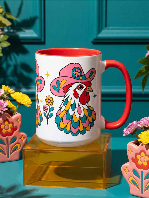Glossy white ceramic coffee mug with a colored rim, inside, and handle and a full wraparound folk art print. Chickens wear pink cowboy hats with teal bands, yellow diamonds, flowers, and teardrop motifs. The birds have white heads, red combs and wattles, and layered pink, teal, and golden yellow feathers outlined in black. Pink and yellow flowers, teal leaves, and four-point starbursts fill the space between. Retro western kitchen decor.