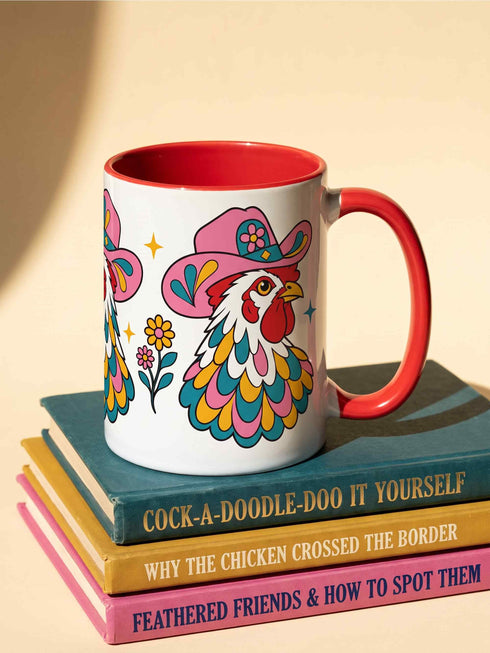 Glossy white ceramic coffee mug with a colored rim, inside, and handle and a full wraparound folk art print. Chickens wear pink cowboy hats with teal bands, yellow diamonds, flowers, and teardrop motifs. The birds have white heads, red combs and wattles, and layered pink, teal, and golden yellow feathers outlined in black. Pink and yellow flowers, teal leaves, and four-point starbursts fill the space between. Retro western kitchen decor.