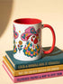 Glossy white ceramic coffee mug with a colored rim, inside, and handle and a full wraparound folk art print. Chickens wear pink cowboy hats with teal bands, yellow diamonds, flowers, and teardrop motifs. The birds have white heads, red combs and wattles, and layered pink, teal, and golden yellow feathers outlined in black. Pink and yellow flowers, teal leaves, and four-point starbursts fill the space between. Retro western kitchen decor.