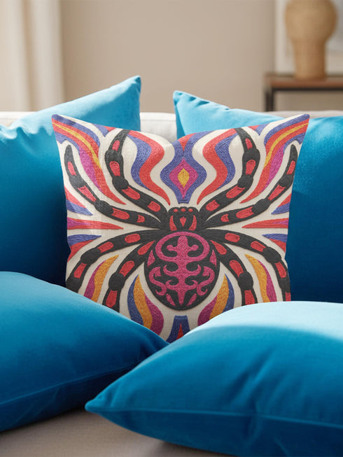 Square psychedelic folk art throw pillow cover printed to look like textured embroidery on a cream background. A large black spider with coral red and magenta markings on its legs and abdomen sits at the center, surrounded by rippling waves in blue violet, coral red, magenta, and golden yellow. Smooth flat 2D print, not raised stitching. Retro gothic Halloween maximalist decor for a sofa, armchair, or goth bedroom, made for spooky season.