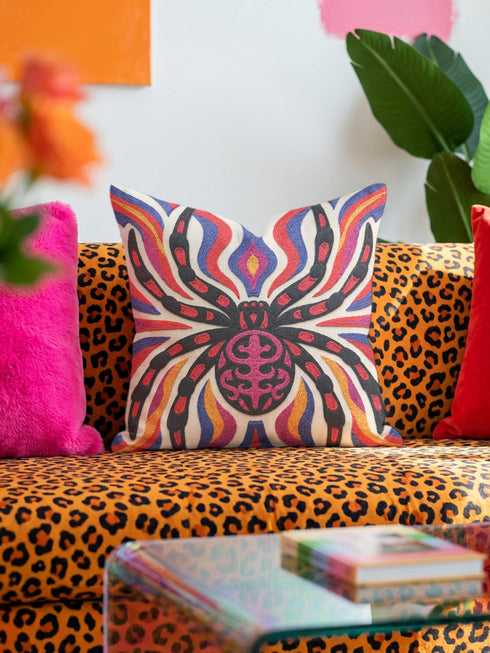 Square psychedelic folk art throw pillow cover printed to look like textured embroidery on a cream background. A large black spider with coral red and magenta markings on its legs and abdomen sits at the center, surrounded by rippling waves in blue violet, coral red, magenta, and golden yellow. Smooth flat 2D print, not raised stitching. Retro gothic Halloween maximalist decor for a sofa, armchair, or goth bedroom, made for spooky season.